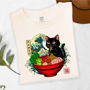 A white t-shirt with a colorful graphic design depicting a black cat sitting in a bowl of ramen noodles, surrounded by waves and vegetables.