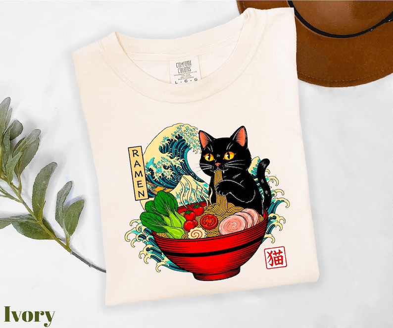 A white t-shirt with a colorful graphic design depicting a black cat sitting in a bowl of ramen noodles, surrounded by waves and vegetables.