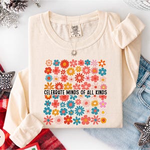 a white long-sleeved t-shirt with a colorful floral design and the text "Celebrate Minds of All Kinds" printed on it. The t-shirt is laid out on a white surface, surrounded by various clothing items such as jeans and a star-shaped embellishment.