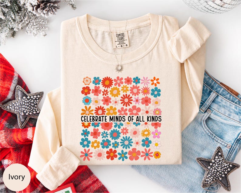a white long-sleeved t-shirt with a colorful floral design and the text "Celebrate Minds of All Kinds" printed on it. The t-shirt is laid out on a white surface, surrounded by various clothing items such as jeans and a star-shaped embellishment.