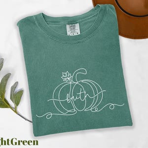 A green t-shirt with a pumpkin and the word "faith" printed on it, along with a brown hat and some greenery, all laid out on a white background.