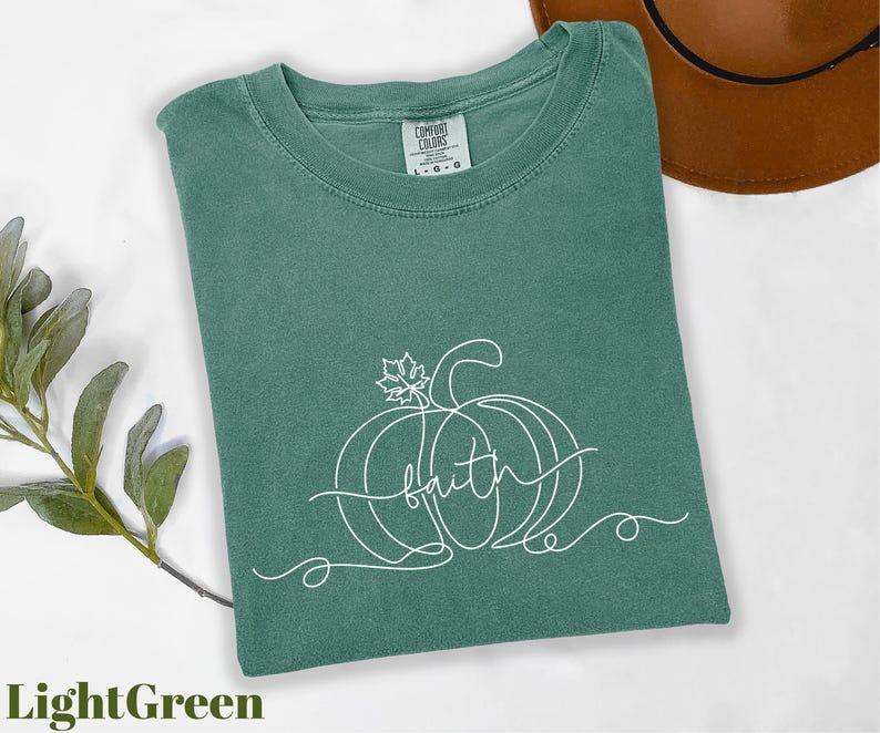 A green t-shirt with a pumpkin and the word "faith" printed on it, along with a brown hat and some greenery, all laid out on a white background.