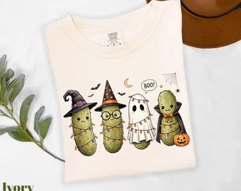 Può includere: Una t-shirt color avorio con una grafica di quattro cetriolini dei cartoni animati vestiti per Halloween. Uno indossa un cappello da strega, uno un cappello da mago con occhiali, uno un lenzuolo da fantasma con luci e un mantello da vampiro.