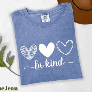A blue t-shirt with the text "be kind" printed on it, along with three heart symbols, is displayed on a white surface.