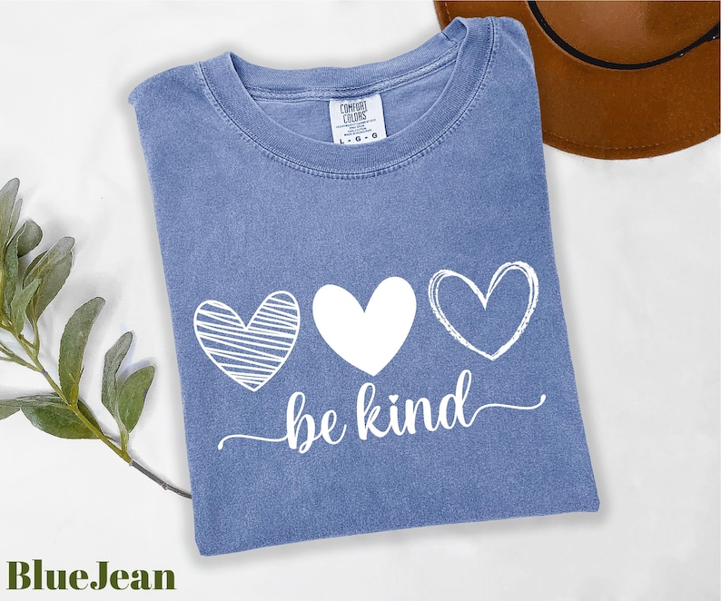A blue t-shirt with the text "be kind" printed on it, along with three heart symbols, is displayed on a white surface.
