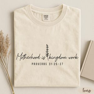 A beige t-shirt with a printed message and a small plant illustration, along with some dried flowers and a book, all placed on a white surface.