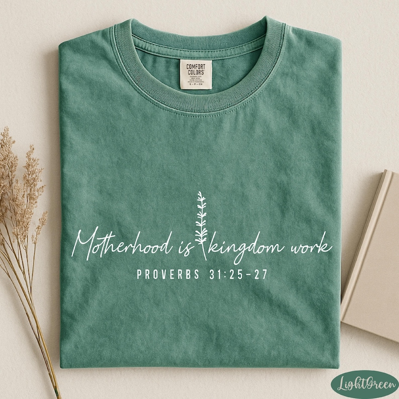 A green t-shirt with white text and a small plant graphic, along with a book and some dried flowers, arranged on a white surface.