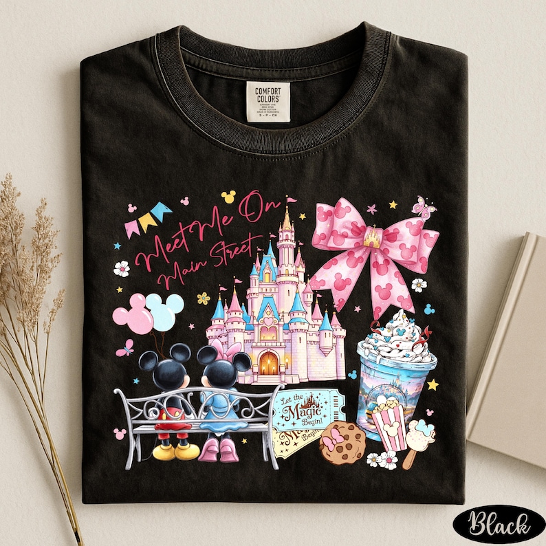 A black t-shirt with a colorful and whimsical design featuring Mickey Mouse, a castle, and various Disney-themed elements such as a cup of ice cream and a cookie.