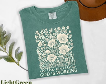 May include: Light green t-shirt with a floral design and the text "in the waiting, God is working." The design features white flowers and foliage. The shirt is made of a soft, comfortable material.