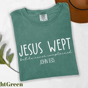 May include: Light green t-shirt with the text "JESUS WEPT but he never complained JOHN 11:35" in white. The shirt is folded and has a Comfort Colors tag. A brown hat is in the background.