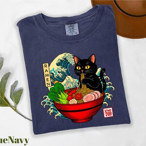 A blue t-shirt with a graphic design depicting a black cat sitting in a bowl of ramen noodles, surrounded by various food items and waves.