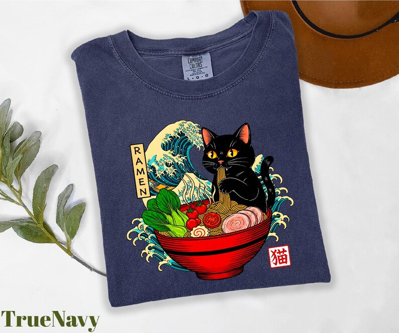 A blue t-shirt with a graphic design depicting a black cat sitting in a bowl of ramen noodles, surrounded by various food items and waves.