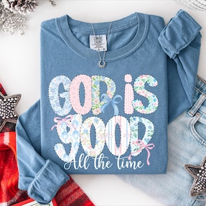 May include: A blue long-sleeve shirt with the words "GOD IS GOOD" in a floral and striped design. The phrase "All the time" is below the main text. A silver necklace and a pair of jeans are next to the shirt.