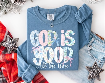 May include: A blue long-sleeve shirt with the words "GOD IS GOOD" in a floral and striped design. The phrase "All the time" is below the main text. A silver necklace and a pair of jeans are next to the shirt.