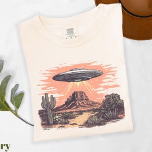 a t - shirt with an image of an alien flying over a desert