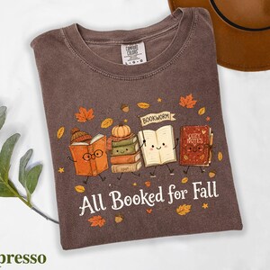 A t-shirt with a graphic design featuring anthropomorphized books and a bookworm, along with the text "All Booked for Fall" and a pumpkin.