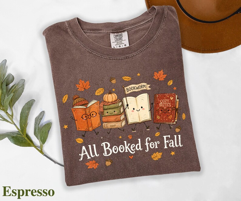 A t-shirt with a graphic design featuring anthropomorphized books and a bookworm, along with the text "All Booked for Fall" and a pumpkin.