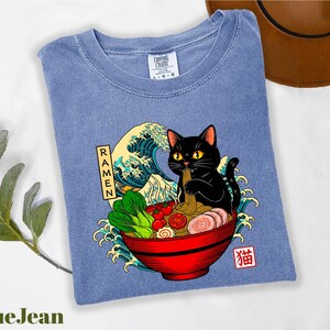 A blue t-shirt with a graphic design depicting a black cat sitting in a bowl of ramen noodles, surrounded by various food items and a wave.