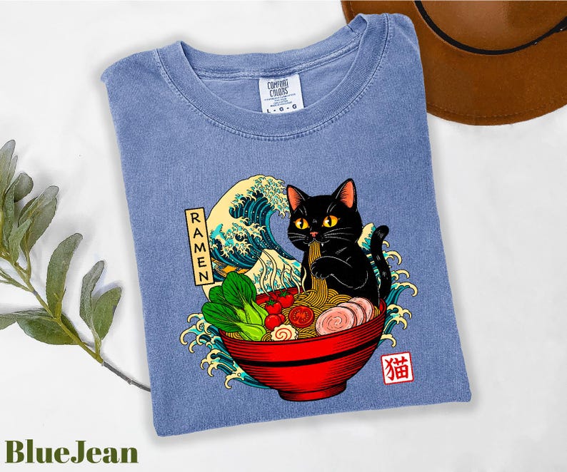 A blue t-shirt with a graphic design depicting a black cat sitting in a bowl of ramen noodles, surrounded by various food items and a wave.