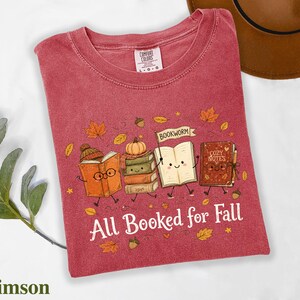 A red t-shirt with a graphic design depicting four anthropomorphic books, each with a unique character, surrounded by autumn leaves and the text "All Booked for Fall".
