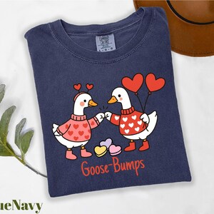A blue t-shirt with a graphic design of two cartoon geese wearing heart-shaped sweaters and holding heart-shaped balloons, with the text "Goose Bumps" below the image.