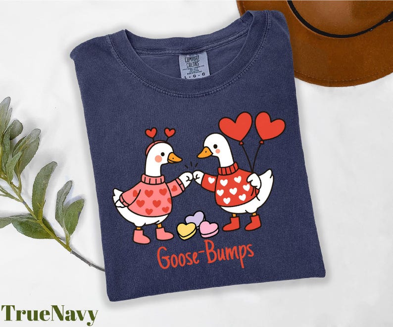 A blue t-shirt with a graphic design of two cartoon geese wearing heart-shaped sweaters and holding heart-shaped balloons, with the text "Goose Bumps" below the image.