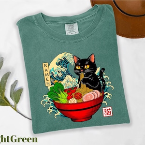 A green t-shirt with a graphic design depicting a black cat sitting in a bowl of ramen noodles, surrounded by various food items and a wave.