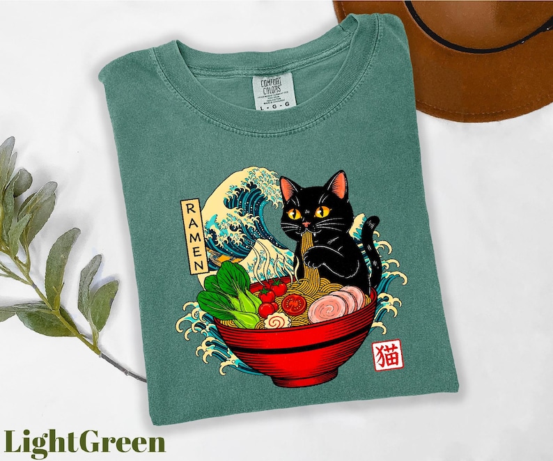 A green t-shirt with a graphic design depicting a black cat sitting in a bowl of ramen noodles, surrounded by various food items and a wave.