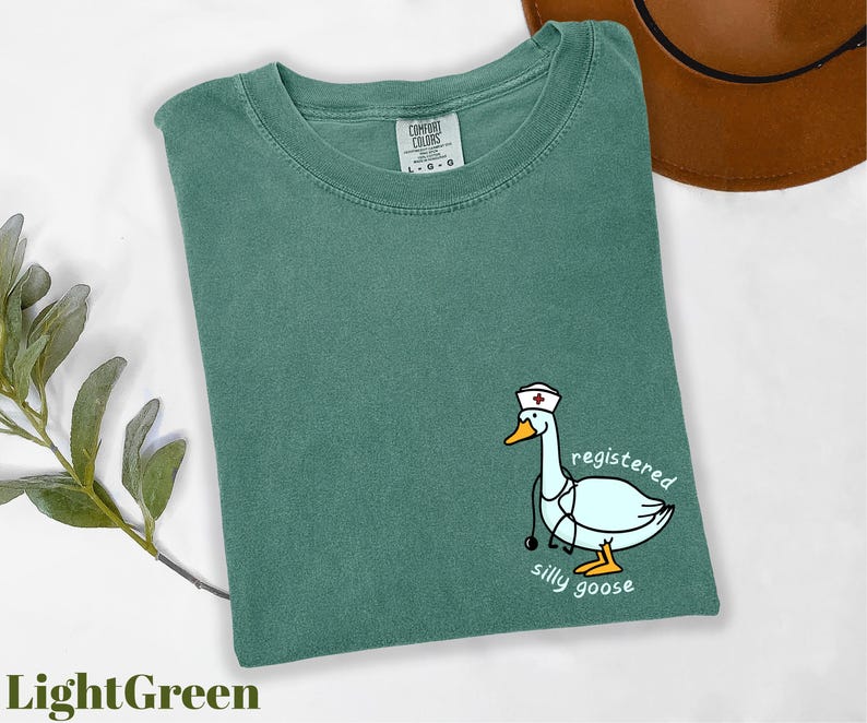 A green t-shirt with a cartoon image of a duck wearing a nurse's hat and the text "registered silly goose" printed on it, along with a brown hat and some greenery.