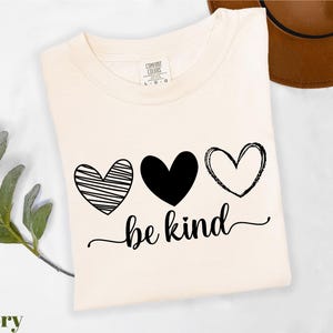 A white t-shirt with a graphic design featuring three hearts and the text "be kind" printed on it, along with a brown hat placed next to it.