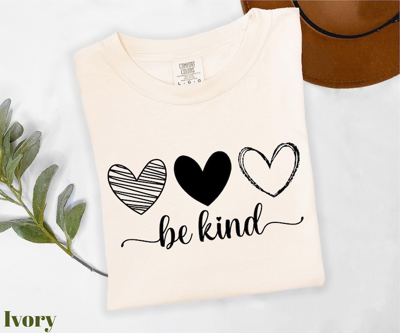 A white t-shirt with a graphic design featuring three hearts and the text "be kind" printed on it, along with a brown hat placed next to it.