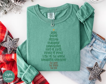 May include: Light green long-sleeve shirt with a Christmas tree design made of text. The text includes the words "Jesus, Savior, Messiah, Redeemer, Emmanuel, King of Kings, Prince of Peace, Light of the World, Wonderful Counselor, The Way, The Truth, The Life."