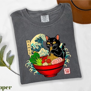 A gray t-shirt with a graphic design depicting a black cat sitting in a bowl of ramen noodles, surrounded by various food items and a wave.