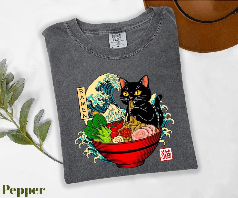 A gray t-shirt with a graphic design depicting a black cat sitting in a bowl of ramen noodles, surrounded by various food items and a wave.