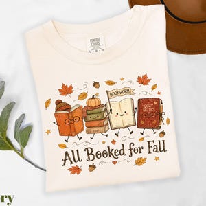 A white t-shirt with a graphic design featuring anthropomorphized books and the text "All Booked for Fall" surrounded by autumn leaves and a pumpkin.