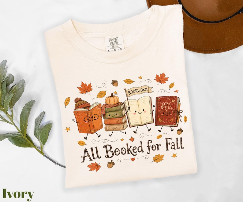 A white t-shirt with a graphic design featuring anthropomorphized books and the text "All Booked for Fall" surrounded by autumn leaves and a pumpkin.