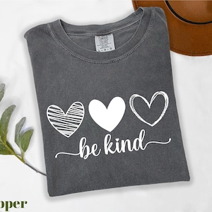 A gray t-shirt with a graphic design of three hearts and the text "be kind" printed on it, along with a brown hat and some greenery.