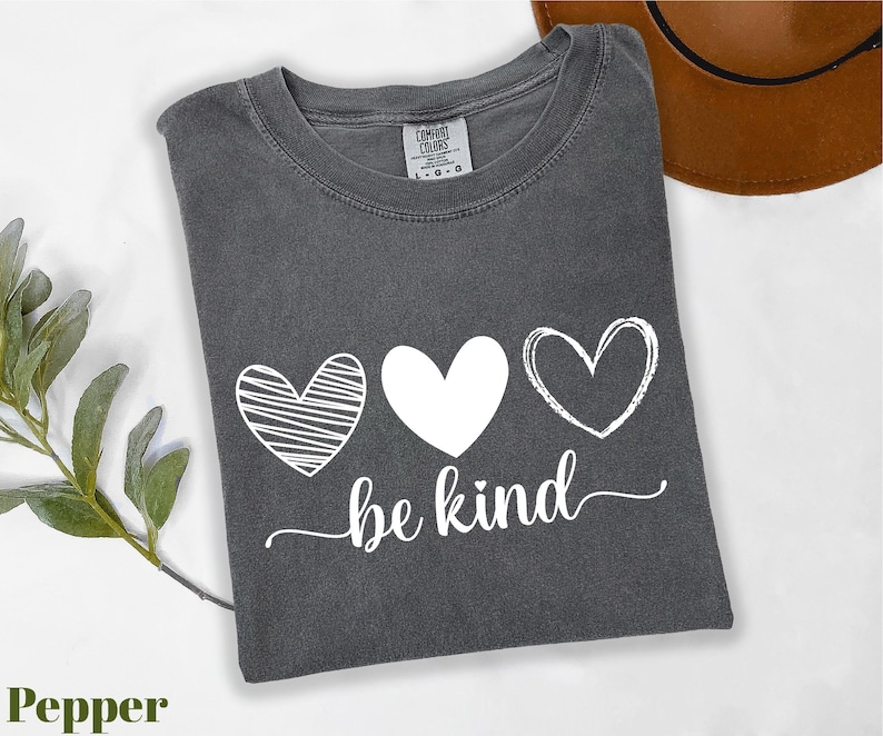 A gray t-shirt with a graphic design of three hearts and the text "be kind" printed on it, along with a brown hat and some greenery.