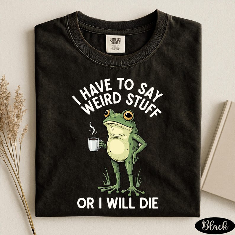a black t-shirt with a humorous graphic of a frog holding a cup of coffee and the text "I HAVE TO SAY WEIRD STUFF OR I WILL DIE" printed on it.
