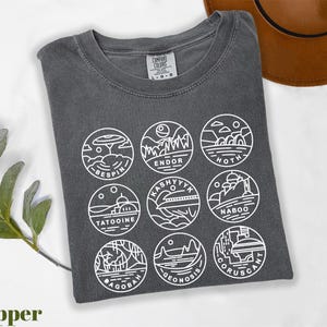 May include: Pepper-colored t-shirt with white line art illustrations of Star Wars planets in circular designs. Planets include Bespin, Endor, Hoth, Tatooine, Kashyyyk, Naboo, Dagobah, Geonosis, and Coruscant. The shirt has a crew neck and short sleeves.