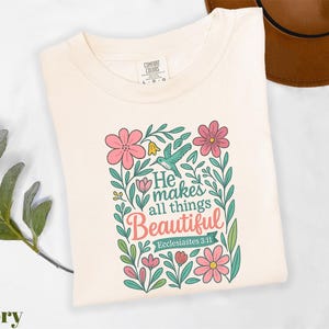 May include: Ivory-colored t-shirt with a floral design and the text "He makes all things Beautiful" from Ecclesiastes 3:11. The design includes pink flowers, green leaves, and a hummingbird.