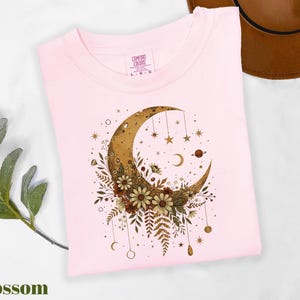 A pink t-shirt with a crescent moon and floral design, along with a brown hat and some greenery, is displayed on a white surface.