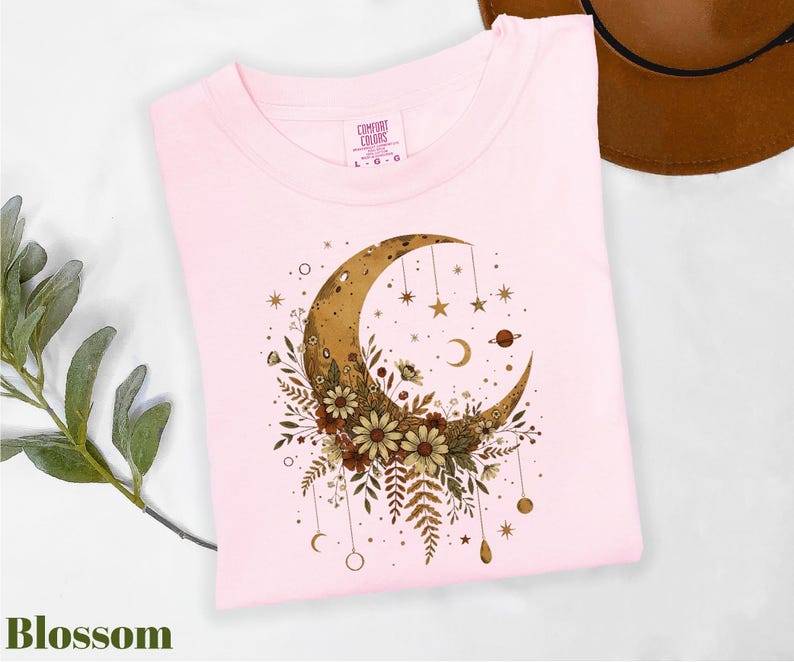 A pink t-shirt with a crescent moon and floral design, along with a brown hat and some greenery, is displayed on a white surface.