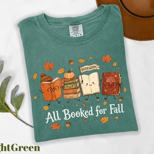 A green t-shirt with a graphic design featuring anthropomorphized books and autumn leaves, along with the text "All Booked for Fall".