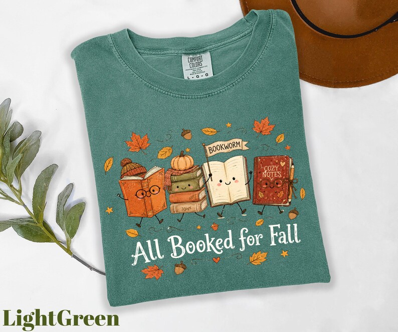 A green t-shirt with a graphic design featuring anthropomorphized books and autumn leaves, along with the text "All Booked for Fall".