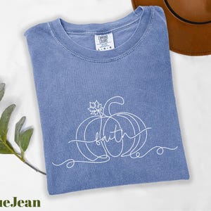 A blue t-shirt with a pumpkin design and the word "Faith" written on it, along with a brown hat and some greenery, are displayed on a white surface.