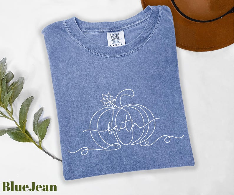 A blue t-shirt with a pumpkin design and the word "Faith" written on it, along with a brown hat and some greenery, are displayed on a white surface.