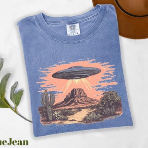 a blue t - shirt with a picture of an alien flying over a desert