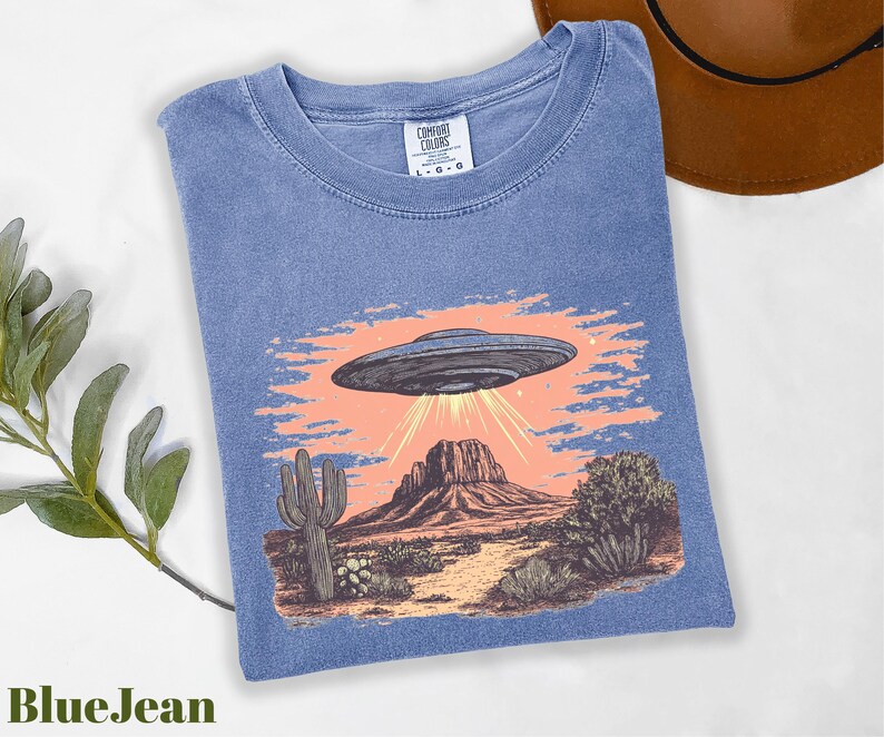 a blue t - shirt with a picture of an alien flying over a desert