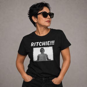 May include: A black short-sleeved t-shirt featuring the white text RITCHIE!!! above a rectangular black and white graphic print of a person looking upward. The shirt has a crew neckline and is made of a solid black fabric with a screen-printed design.